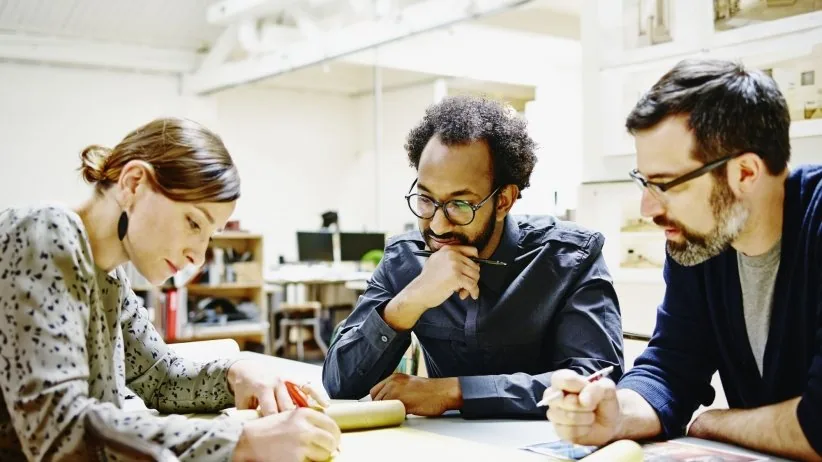 Three colleagues leaning over a workbench together, sketching and talking through an idea.
