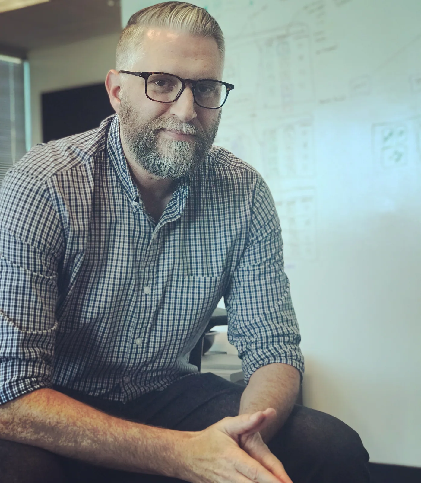 Jeff sitting on a desk in front of a whiteboard covered in system diagrams, wearing a checked shirt and glasses.