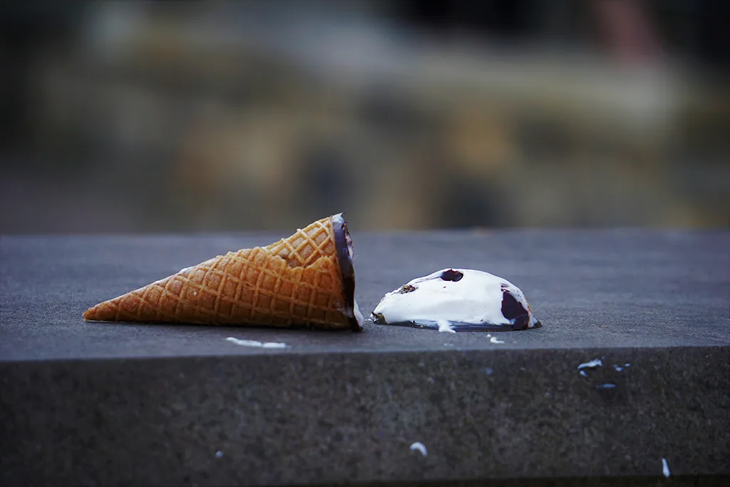 A dropped ice cream cone lying on a stone ledge next to its fallen scoop.