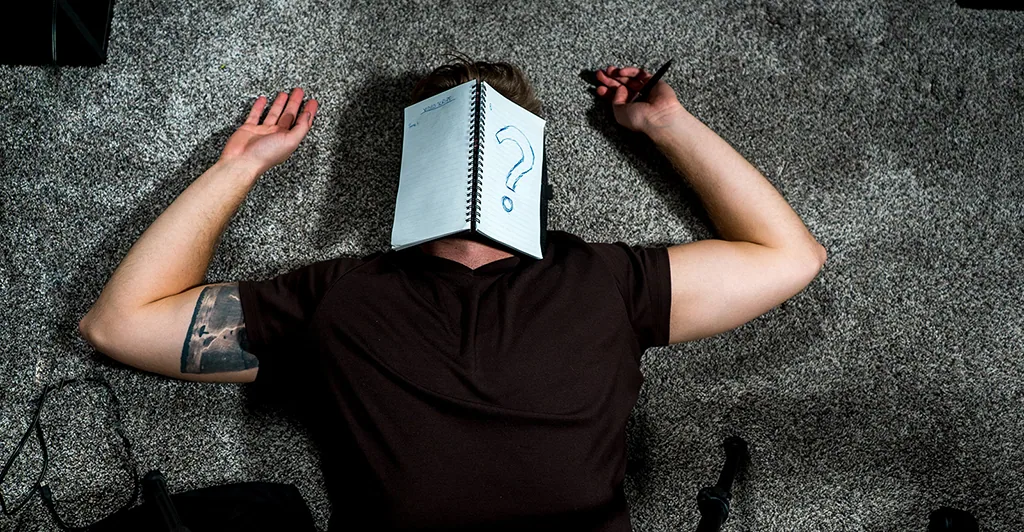 A person lying face-up on the carpet with an open notebook covering their face, a large question mark drawn on the page.